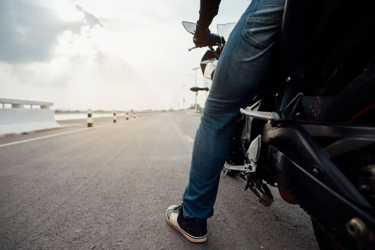Na imagem, as pernas de um homem em cima de uma moto, parado em uma rua