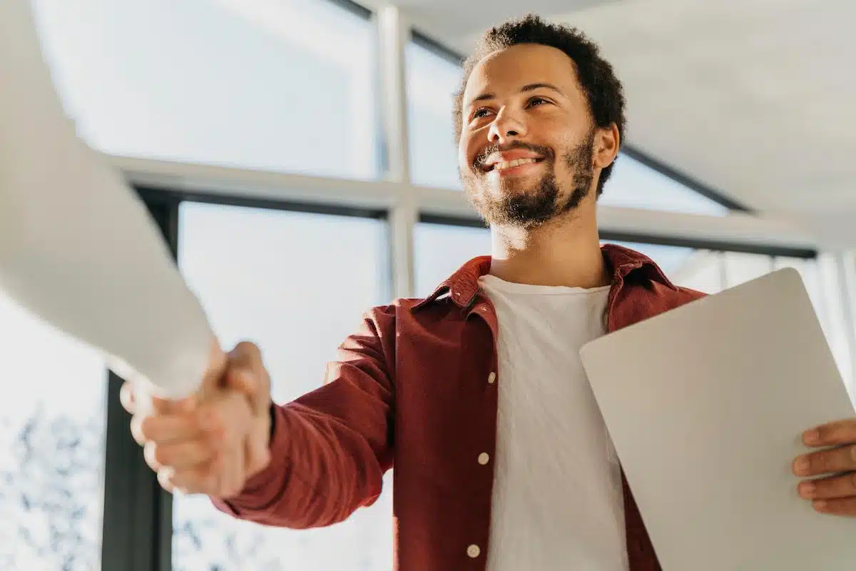Foto mostra duas pessoas apertando as mãos, uma à frente da imagem, segurando um papel. Representação de uma contratação para emprego.