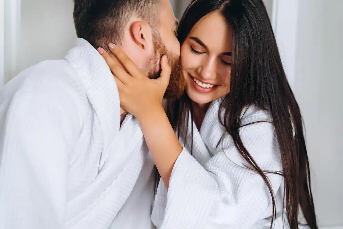 Foto mostra pessoas vestindo roupão branco. Um homem inclinado beijando uma mulher no rosto. Imagem ilustrativa da compatibilidade entre signos.