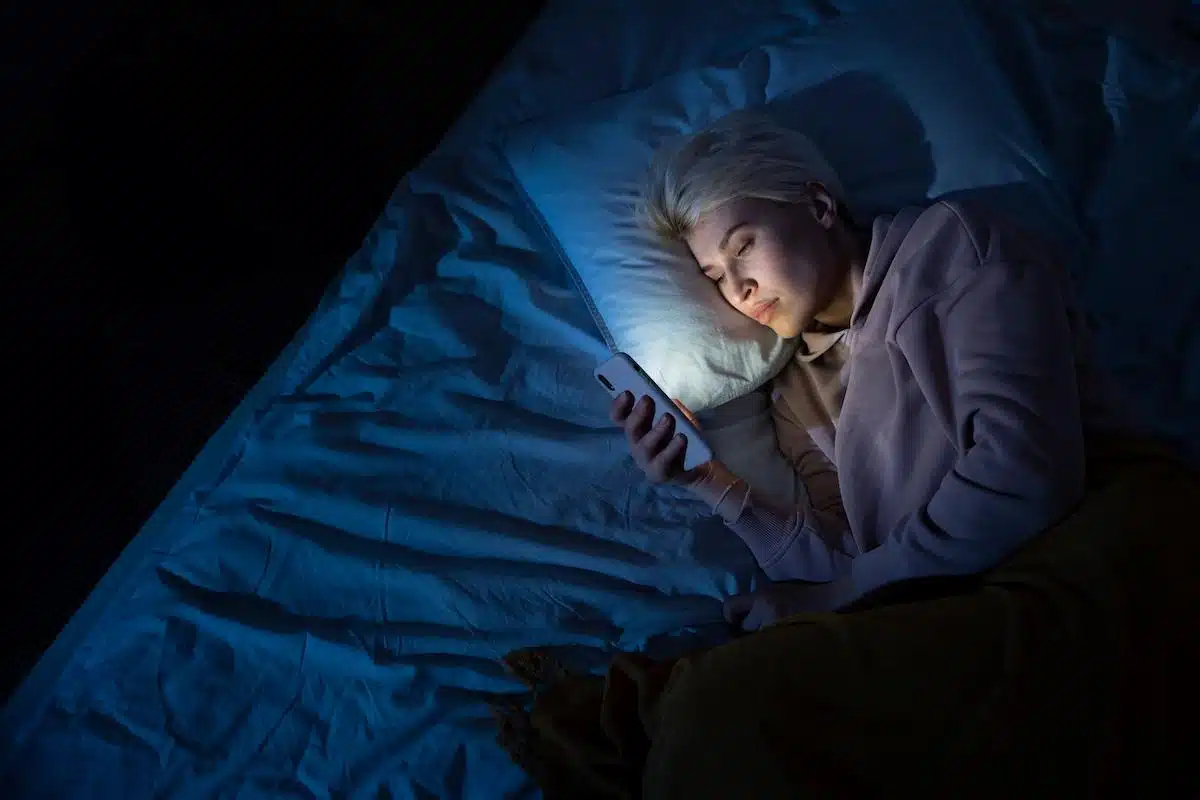 Foto mostra mulher usando celular, deitada em cama em um quarto escuro. Imagem representativa de insônia às 3 horas.