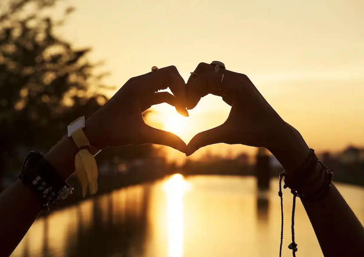 Foto mostra duas mãos fazendo gesto de amor, em um pôr do sol. Imagem ilustrativa do amor que pode ser encontrado por pessoas de certos signos.