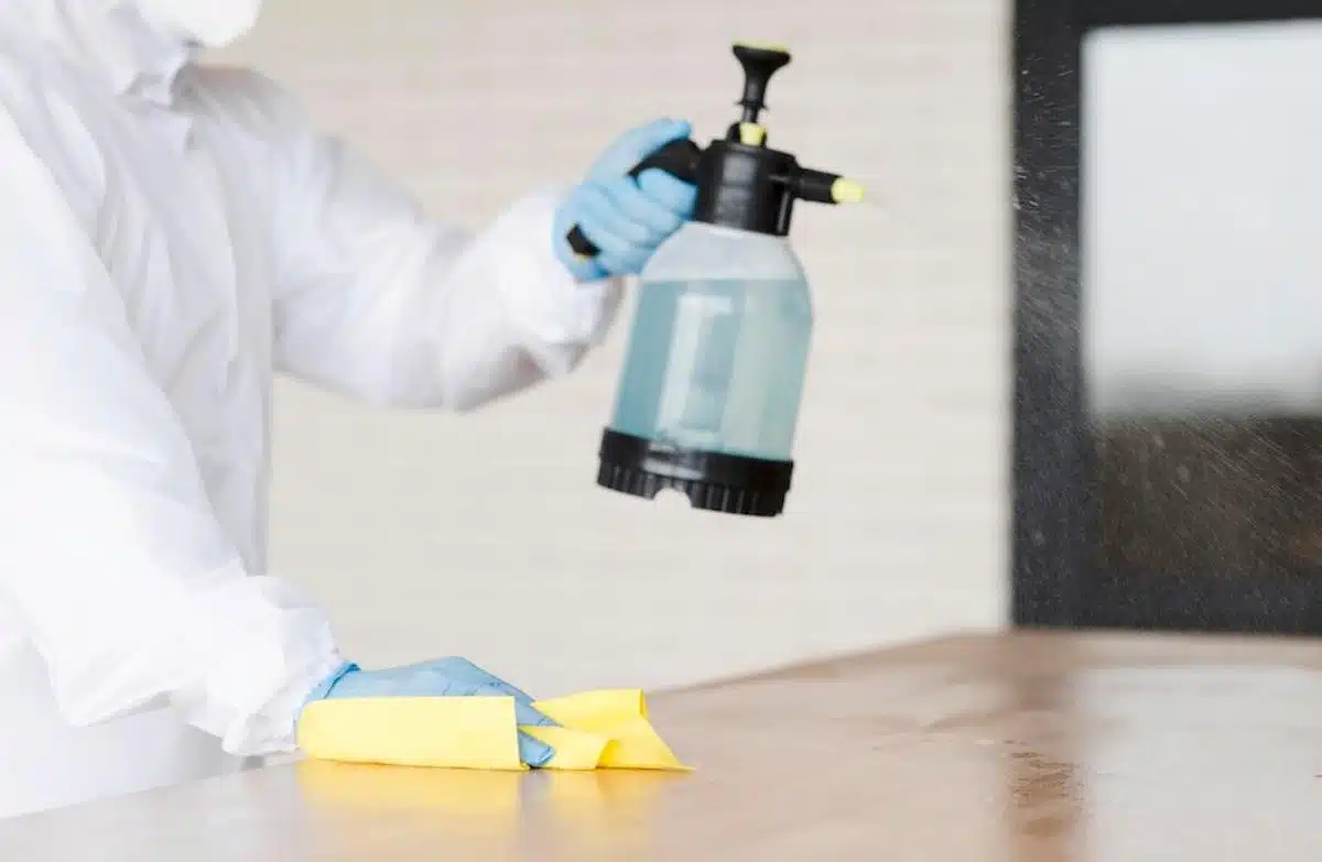 Foto mostra pessoa usando equipamentos de proteção sanitária, borrifando um tipo de produto azulado em um ambiente doméstico, enquanto segura um pano amarelo em sua mão, apoiada acima de uma superfície. Imagem ilustrativa dos odores que afastam baratas.