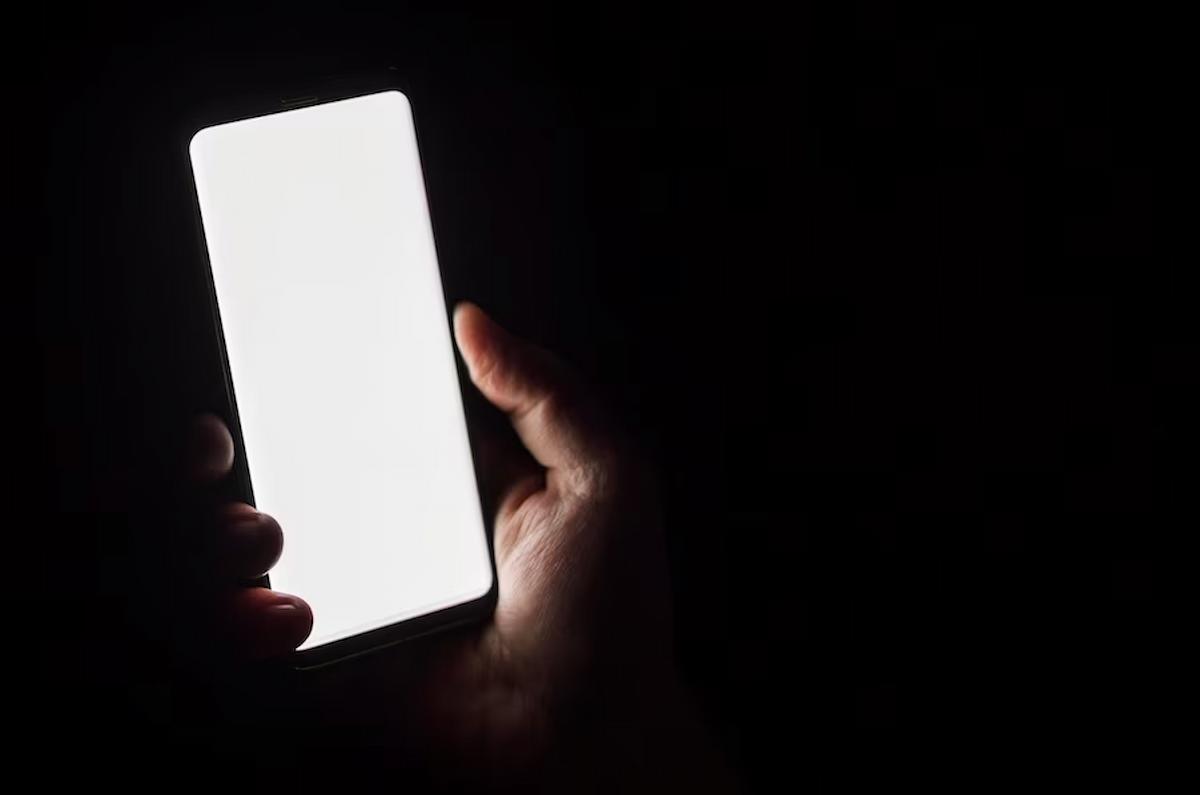 Foto mostra uma mão de uma pessoa usando um celular em um ambiente escuro.