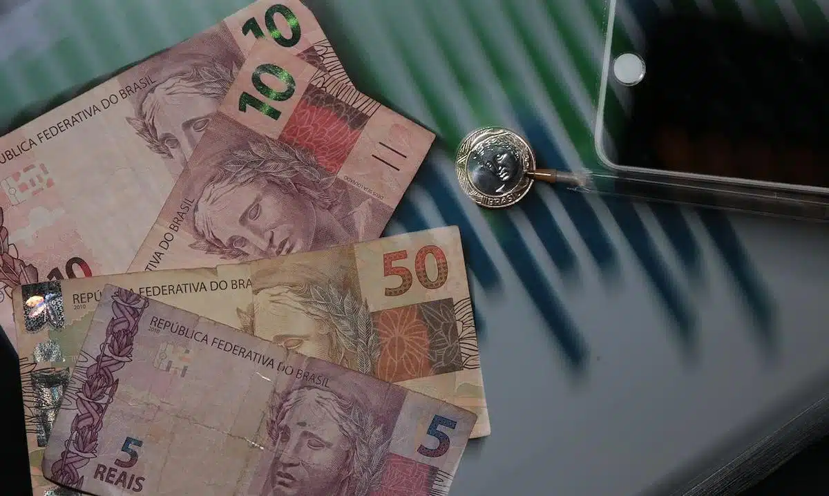 Foto mostra diversas cédulas de real, com notas de R$ 50, R$ 10 e R$ 5, além de uma moeda. Todas estão acima de uma superfície, ao lado de uma caneta e um celular.