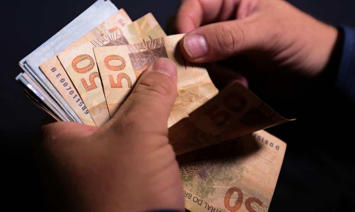 Foto mostra pessoa segurando cédulas de real, com o objetivo de representar o pagamento do 13º salário.