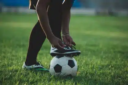 Imagem mostra pessoa amarrando chuteira com o é apoiado em bola de futebol.