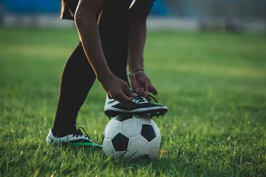 Imagem mostra pessoa amarrando chuteira com o é apoiado em bola de futebol.