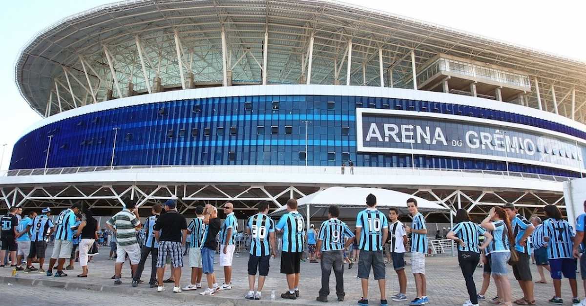 Foto mostra o estádio do Grêmio, com uma fila de torcedores à frente.
