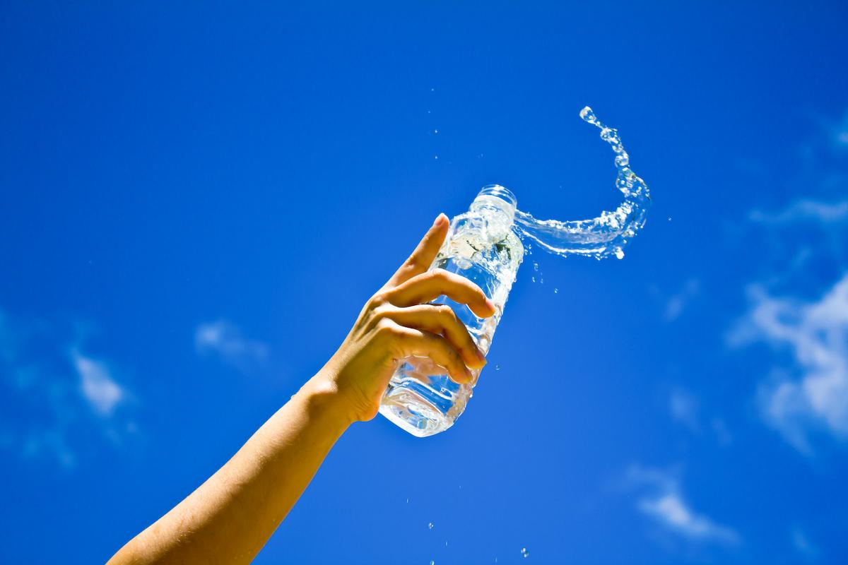 Foto mostra pessoa segurando garrafa de água em direção ao céu.