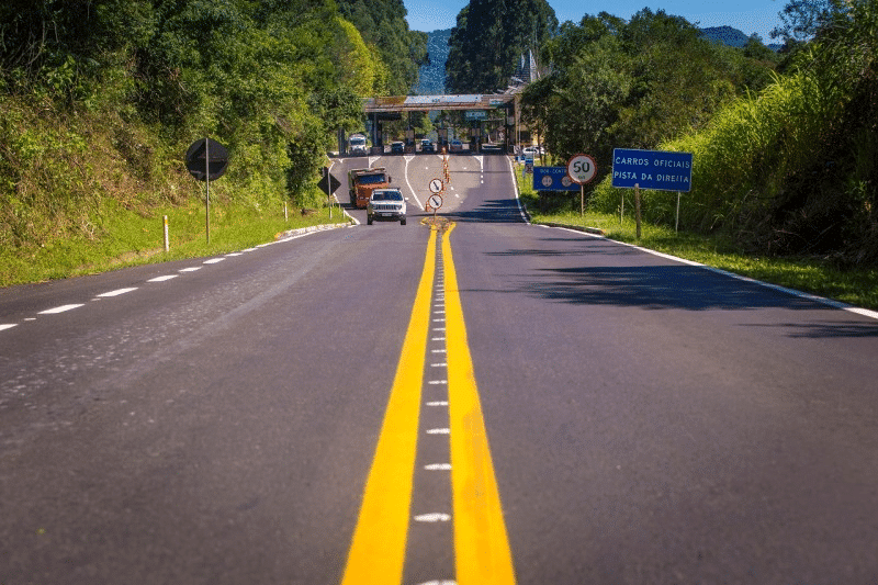 Foto mostra trânsito em rodovia gaúcha, com vegetação ao lado e um posto de pedágio ao fundo da imagem.