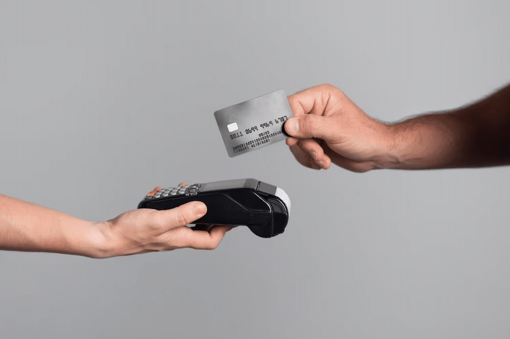 Foto mostra mão de duas pessoas, uma segurando um cartão de crédito, e a outra ao lado oposto, segurando uma maquininha, representando uma transação.