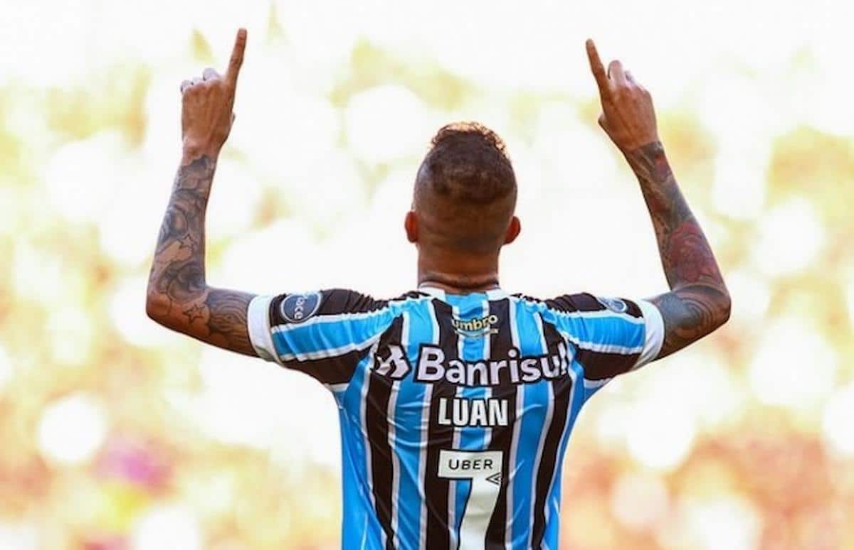 Foto mostra o atleta Luan, em campo com a camisa do Grêmio, levantando os braços para cima em gesto de comemoração ou agradecimento.