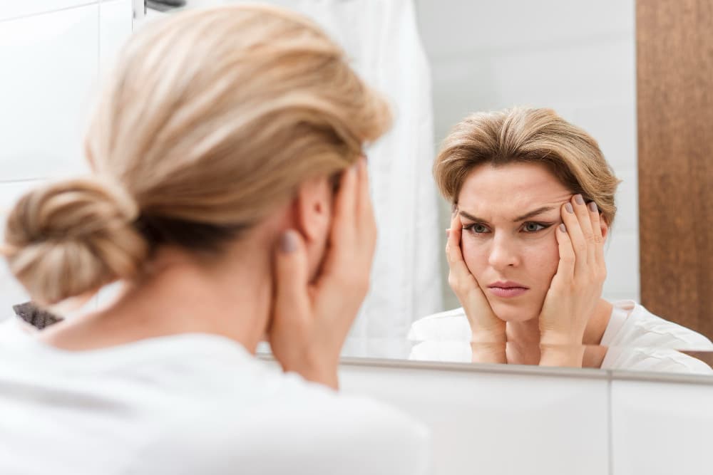 Na imagem, uma mulher loira segurando o rosto na frente do espelho
