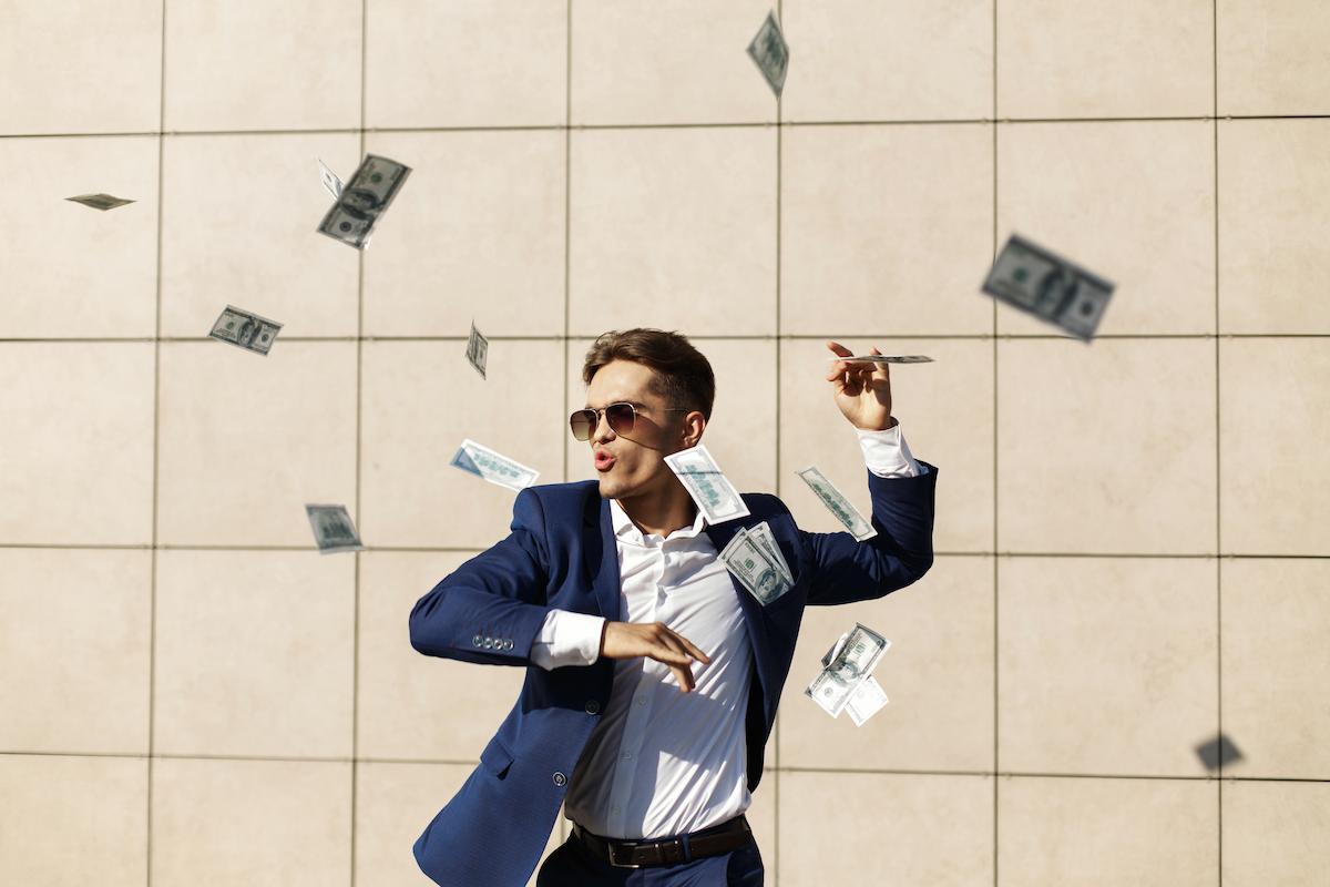 Foto mostra homem com roupas formais celebrando enquanto cédulas de dinheiro caem ao seu redor. Imagem ilustrativa do dinheiro extra que pode ser recebido por alguns signos no mês de julho.