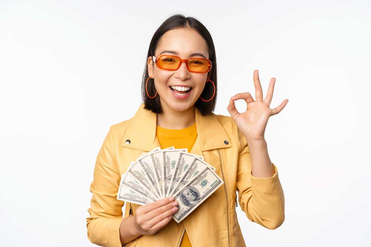 Foto mostra mulher vestida de amarelo, com óculos, segurando cédulas de dinheiro, fazendo gesto de "ok" com a outra mão. Imagem ilustrativa das simpatias que podem atrair dinheiro.
