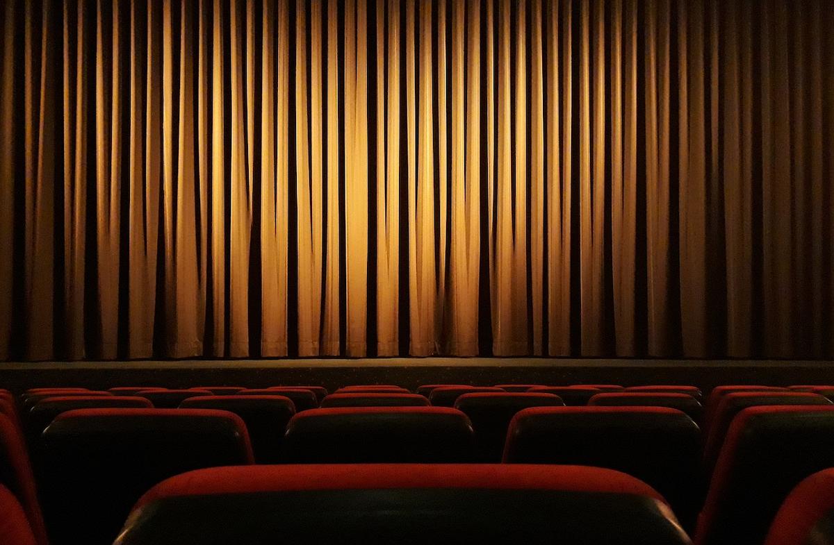 Foto mostra teatro, mostrando cortina sob o palco e cadeiras a frente.