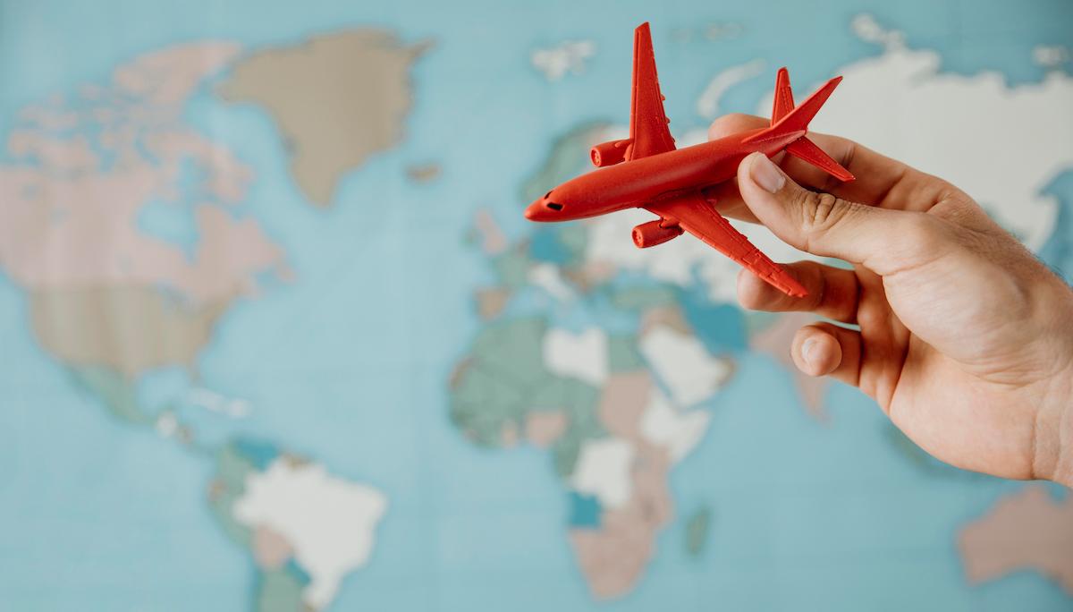 Foto representa voo com pessoa segurando avião a cima de um mapa mundi.